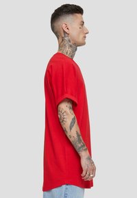 Camiseta de algodón roja con mangas cortas, corte relajado, escote redondo y costuras visibles. El modelo posa de lado, mostrando tatuajes en el brazo y el cuello.