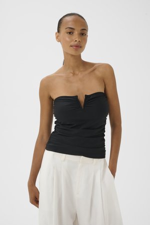 Femme portant un haut noir bustier avec une encoche en V et un pantalon blanc ample, debout devant un fond blanc uni.