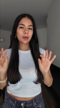 Mladá žena s dlhými tmavými vlasmi, oblečená v bielej krátkej košeli a džínsoch, gestikuluje oboma rukami v interiéri.