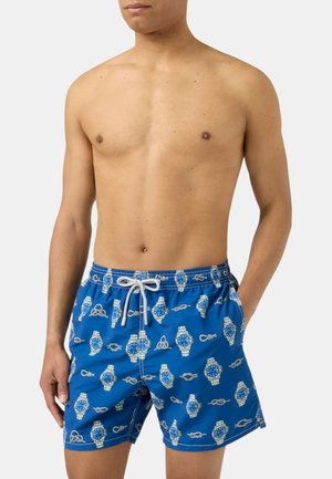 Torace maschile con pantaloncini da bagno blu con motivi di orologi e nodi, mani in tasca, su uno sfondo chiaro uniforme.