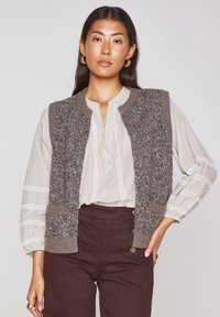 Gilet grigio texturizzato con colletto a coste e tasche, indossato sopra una camicia bianca a righe con bottoni. I pantaloni marroni a vita alta completano il look.