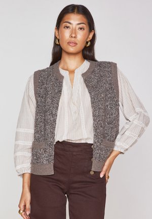 Gray textured vest with a ribbed collar and pockets, layered over a white striped button-up shirt. Brown high-waisted pants complete the look.