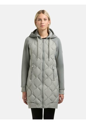 Kobieta ubrana w jasno szarą, pikowaną bluzę z kapturem zapinaną na zamek błyskawiczny, z kieszeniami na zamek z przodu i długimi rękawami, stojąca na jednolitym tle.