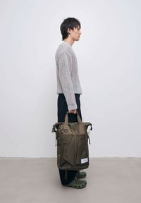 Olivevihreä reppu, joka on valmistettu kestävästä materiaalista, verkkomaisilla etutaskuilla, kaksilla kahvoilla ja vetoketjukiinnityksellä. Mallilla on päällä harmaa neulepusero ja mustat housut.