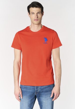 Uomo che indossa una t-shirt arancione a maniche corte con il logo USPA blu sul petto e jeans blu, in piedi davanti a uno sfondo chiaro e uniforme.