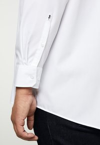 Witte blouse met lange mouwen en geknoopte manchetten, voorzien van een geborduurd zwart bloemenpatroon op de mouwen. Glad materiaal en een getailleerde pasvorm.