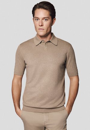 Koszulka polo z krótkim rękawem w kolorze beżowym, wykonana z miękkiego materiału. Posiada kołnierzyk, zapinanie na guziki i ściągacz u dołu. Noszona z pasującymi spodniami.