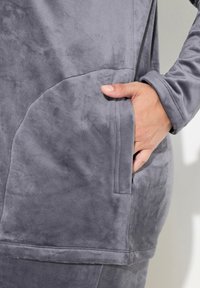 Tunic in tessuto plush grigio e morbido con tasca laterale. Presenta un design semplice e polsini a costine, mettendo in risalto la sua texture liscia.