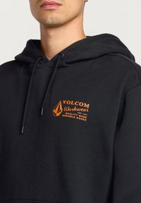 Sort hættejakke lavet af bomuld, med et frontlogo i orange. Designet inkluderer en hætte med snore og ribbede manchetter. Holdbart stof er synligt.