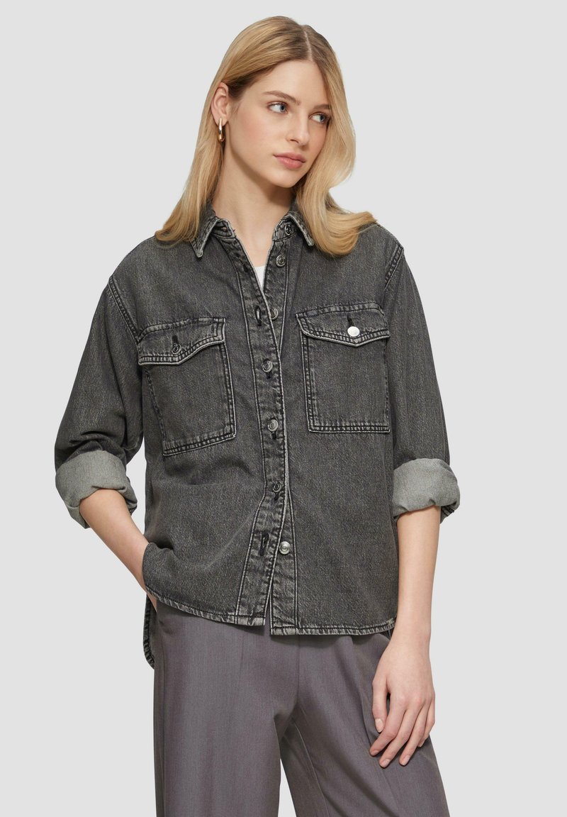 Czarna koszula dżinsowa z dwiema kieszeniami piersiowymi, zapinana na guziki z podwiniętymi rękawami, w zestawie z szarymi spodniami. Gładka tekstura i klasyczny design.