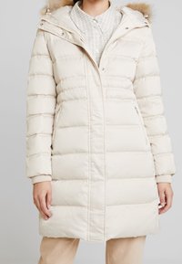 Beige padded long coat with hood, fur trim, and ribbed cuffs. Features a zip front and two side pockets. Soft, smooth texture.