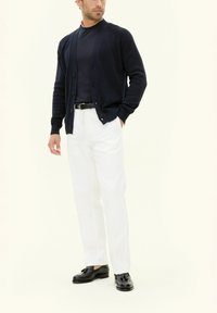 Mörkblå tröja med knäppning över mörk skjorta, vita byxor och svarta skor. Har ribbade ärmslut och knappar, med en polerad och välklädd design.