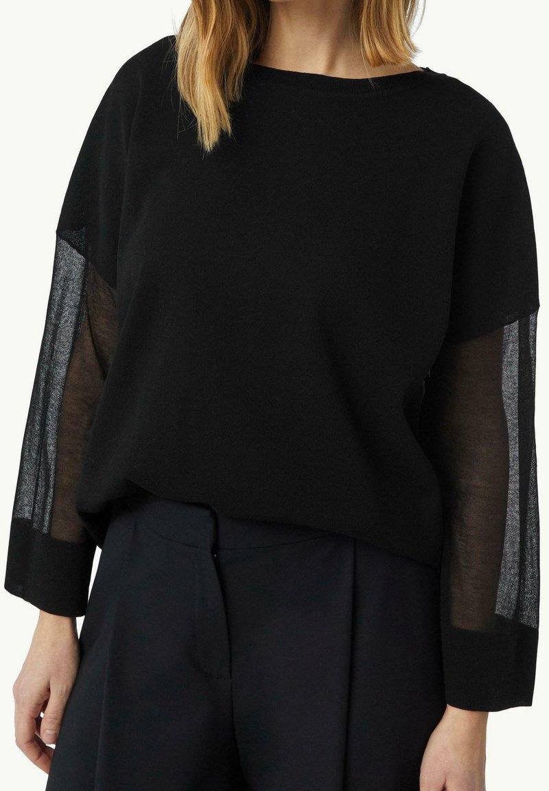 Femme portant un haut noir à manches longues avec des panneaux en tissu transparent sur les manches et un pantalon noir taille haute sur fond blanc.