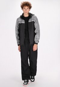 Giacca a vento nera e grigia con zip frontale, cappuccio e tasche laterali, abbinata a pantaloni neri larghi e sneakers nere con dettagli bianchi.