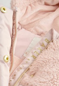 Rosa jacka med en plyschig, fluffig inredning. Har en guldfärgad dragkedja och detaljer i guld. Etikett med präglat text synlig.
