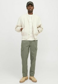 Light beige bomber jacket over a white t-shirt, paired with olive green cargo pants. Tan boots complete the outfit. Cap in black.
