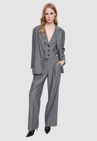 Donna che indossa un completo grigio a tre pezzi con blazer, gilet abbottonato, pantaloni a gamba larga e scarpe nere a punta, in piedi con una mano in tasca.