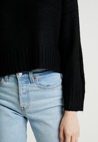 Czarny dzianinowy sweter z prążkowanym dołem, noszony na jasnoniebieskich dżinsach. Sweter ma opadające ramiona i fakturowany materiał.