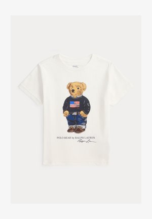 Biela košeľa s krátkym rukávom, na ktorej je medveď v svetri s americkou vlajkou, džínsy a topánky, označené "Polo Bear od Ralph Lauren."