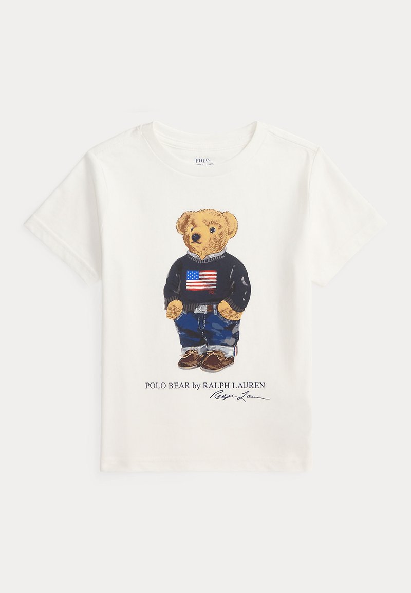 Biela košeľa s krátkym rukávom, na ktorej je medveď v svetri s americkou vlajkou, džínsy a topánky, označené "Polo Bear od Ralph Lauren."