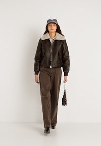 Smeđa kožna bomber jakna s krznom na ovratniku, u paru s širokim smeškim hlačama i sivoj kapom. Outfiti uključuje malu torbicu.