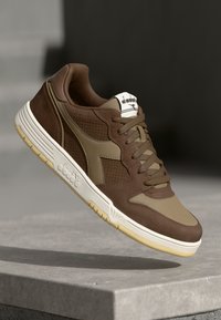 Brown athletic shoes featuring a mix of smooth leather and textured fabric, with a cream sole and subtle logo accents on the tongue and side.