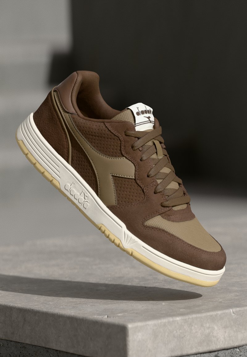 Brown athletic shoes featuring a mix of smooth leather and textured fabric, with a cream sole and subtle logo accents on the tongue and side.
