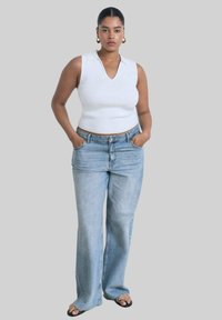 Vit ärmlös topp med krage, ihop med ljusblå vida jeans. Modellen står med händerna i fickorna och bär svarta sandaler med öppen tå.