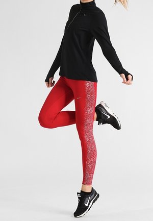Top deportivo de manga larga negro con cuello con cremallera, combinado con leggings rojos que presentan un patrón abstracto texturizado, y zapatillas negras con detalles blancos.