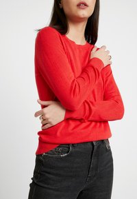 Maglione rosso a maniche lunghe con scollatura rotonda e polsini a coste, abbinato a jeans attillati grigio scuro. Tessuto morbido e testurizzato con una vestibilità comoda.