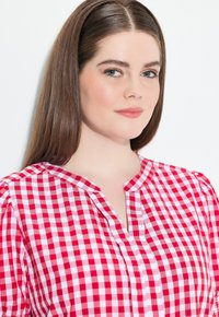 Blouse en vichy rouge et blanc avec un col en V, des manches courtes bouffantes et de subtils plis. Fabriquée en tissu léger avec un motif à carreaux.