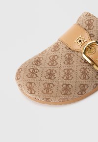 Tan instapschoen met geperst monogrampatroon, beige leren band, gouden gesp en decoratief gouden embleem op witte achtergrond.