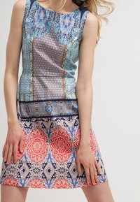 Šaty bez rukávů s modrým vrškem s detailními vzory a rozšířenou sukní s geometrickými a květinovými vzory v červené, bílé a modré.