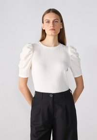 Top alb texturat cu mâneci scurte tip puff, asociat cu pantaloni negri cu un nasture în față. Material neted și design ajustat.