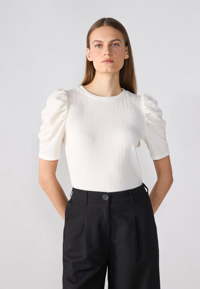 Top alb texturat cu mâneci scurte tip puff, asociat cu pantaloni negri cu un nasture în față. Material neted și design ajustat.