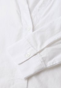 Camisa blanca de algodón con cuello puntiagudo, que presenta una textura suave, detalles de botones y mangas largas. Patrón diagonal sutil visible.