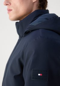 Marineblå hættetrøje i struktureret stof, med et logo-patch på skulderen. Designet inkluderer en høj krave og kontrasterende mønstre.