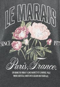 Tela gris oscuro con texto blanco y diseño floral. Peonías en tonos rosa y crema con hojas verdes. El texto incluye "LE MARAIS", "París, Francia" y "Desde 1977".