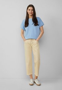 Top azzurro chiaro a maniche corte, taglio morbido. Pantaloni crema, corti con orlo sfrangiato. Sneakers bianche e beige. Sfondo semplice.