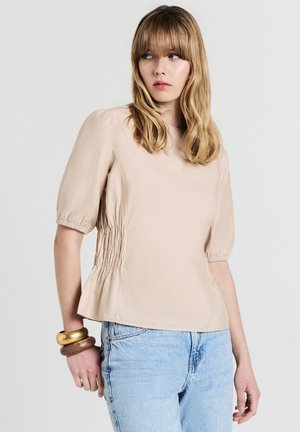 Blusa beige a maniche corte con scollatura rotonda, caratterizzata da arricciature sui lati. Indossata con jeans in denim blu chiaro e braccialetti sovrapposti.
