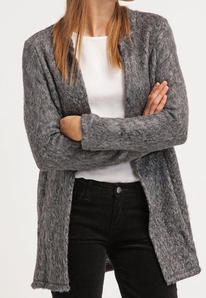 Person trägt eine strukturierte graue Strickjacke mit offenem Vorderteil über einem weißen Hemd und schwarzen Hosen, die Arme vor der Brust verschränkt.