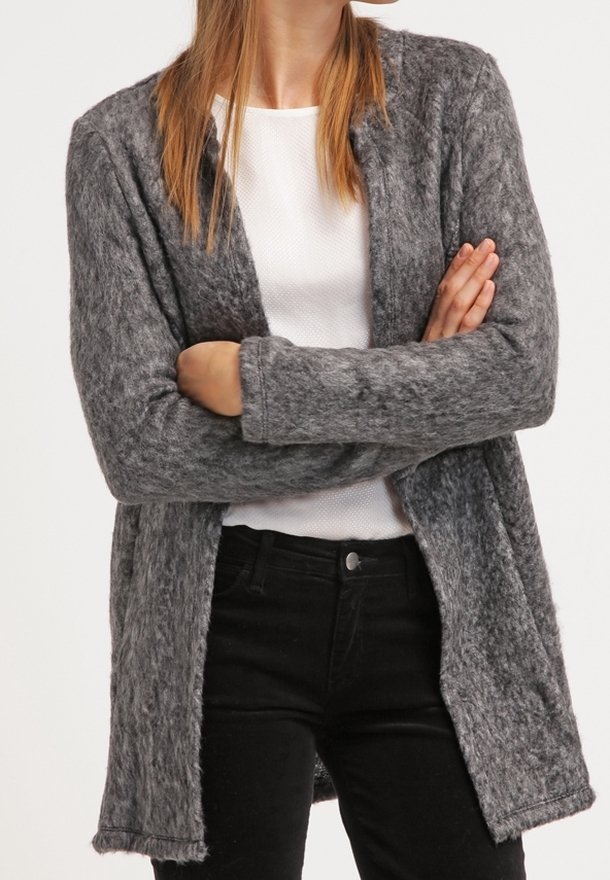 Personne portant un cardigan gris texturé à ouverture sur le devant, par-dessus une chemise blanche et un pantalon noir, les bras croisés sur la poitrine.
