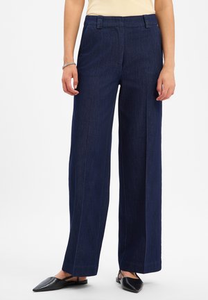 Femme portant un pantalon large bleu foncé, un haut beige, un bracelet argenté et des chaussures plates noires pointues sur fond blanc.
