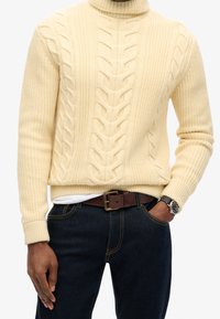 Pull à col roulé crème en maille torsadée avec motif texturé, associé à un jean foncé et une ceinture marron. Montre visible au poignet.