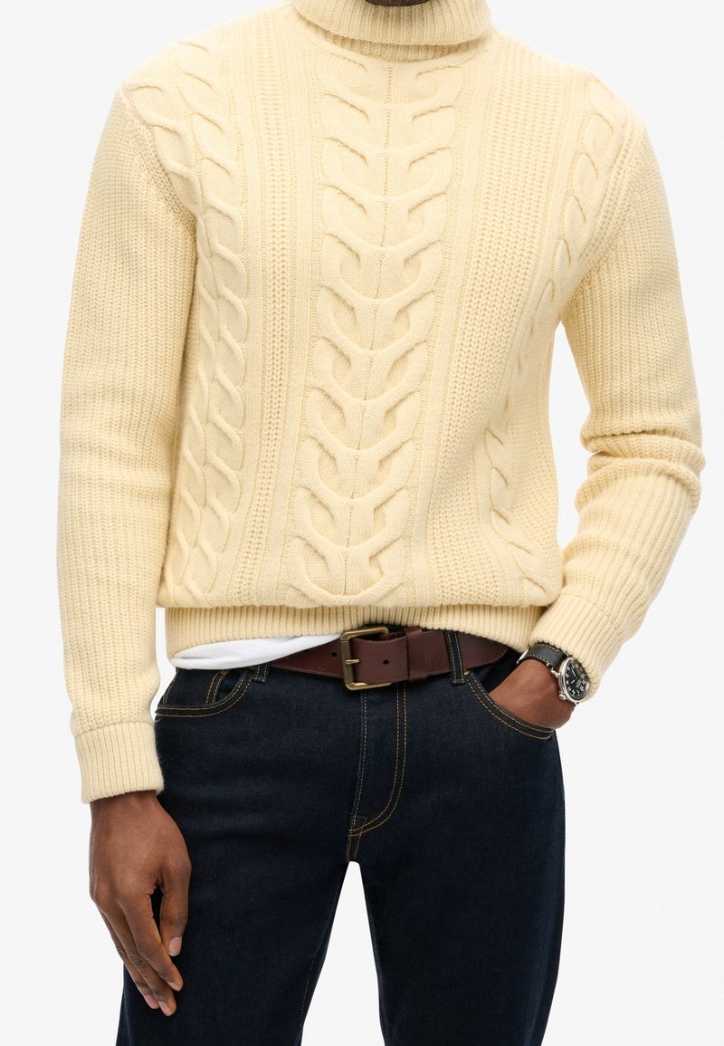 Pull à col roulé crème en maille torsadée avec motif texturé, associé à un jean foncé et une ceinture marron. Montre visible au poignet.