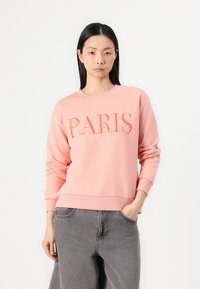 Rózsaszín pulóver, amelyen narancssárga hímzett "PARIS" felirat található. Bordázott mandzsettákkal és aljjal rendelkezik, szürke, bő szabású nadrággal viselve, sima textúrával.
