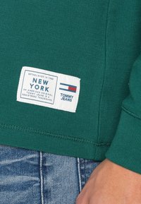 Zelený žebrovaný svetr s bílou značkovou nášivkou. Nášivka obsahuje nápisy "NEW YORK" a "TOMMY JEANS" s logovými pruhy v červené a modré barvě.