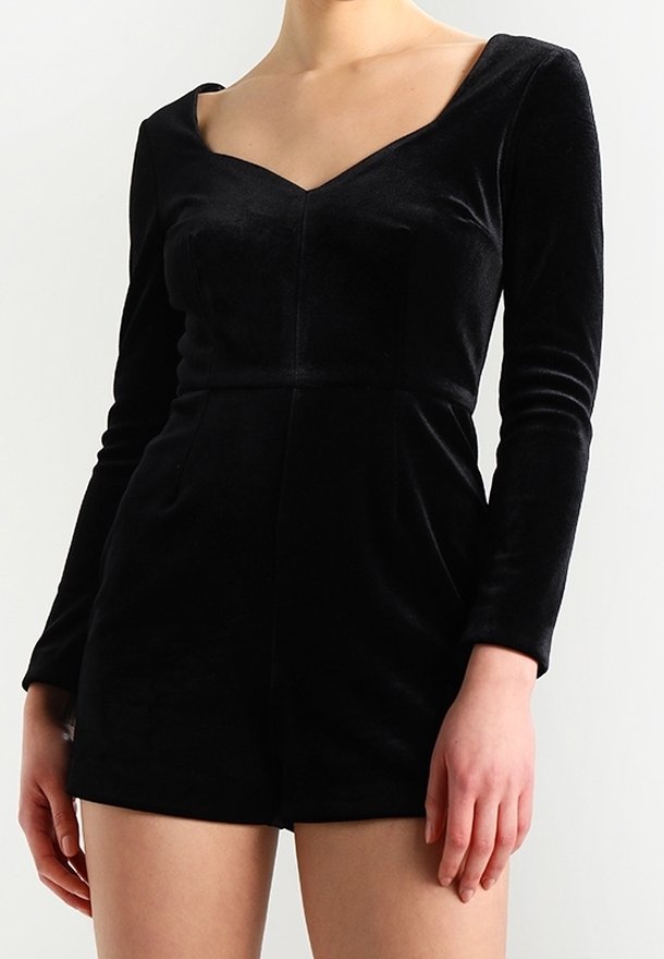 Femme portant une combinaison-short en velours noire à manches longues, avec un décolleté carré plongeant et une taille cintrée.