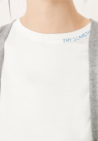 Camiseta de algodón blanca con cuello redondo y texto bordado azul "PRUEBA ALGO" en el hombro, combinada con un cárdigan gris claro.