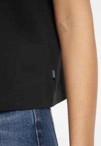 T-shirt en coton noir exposé, avec une étiquette latérale portant la mention "GUESS". Le design est simple avec un ourlet droit et des manches courtes.